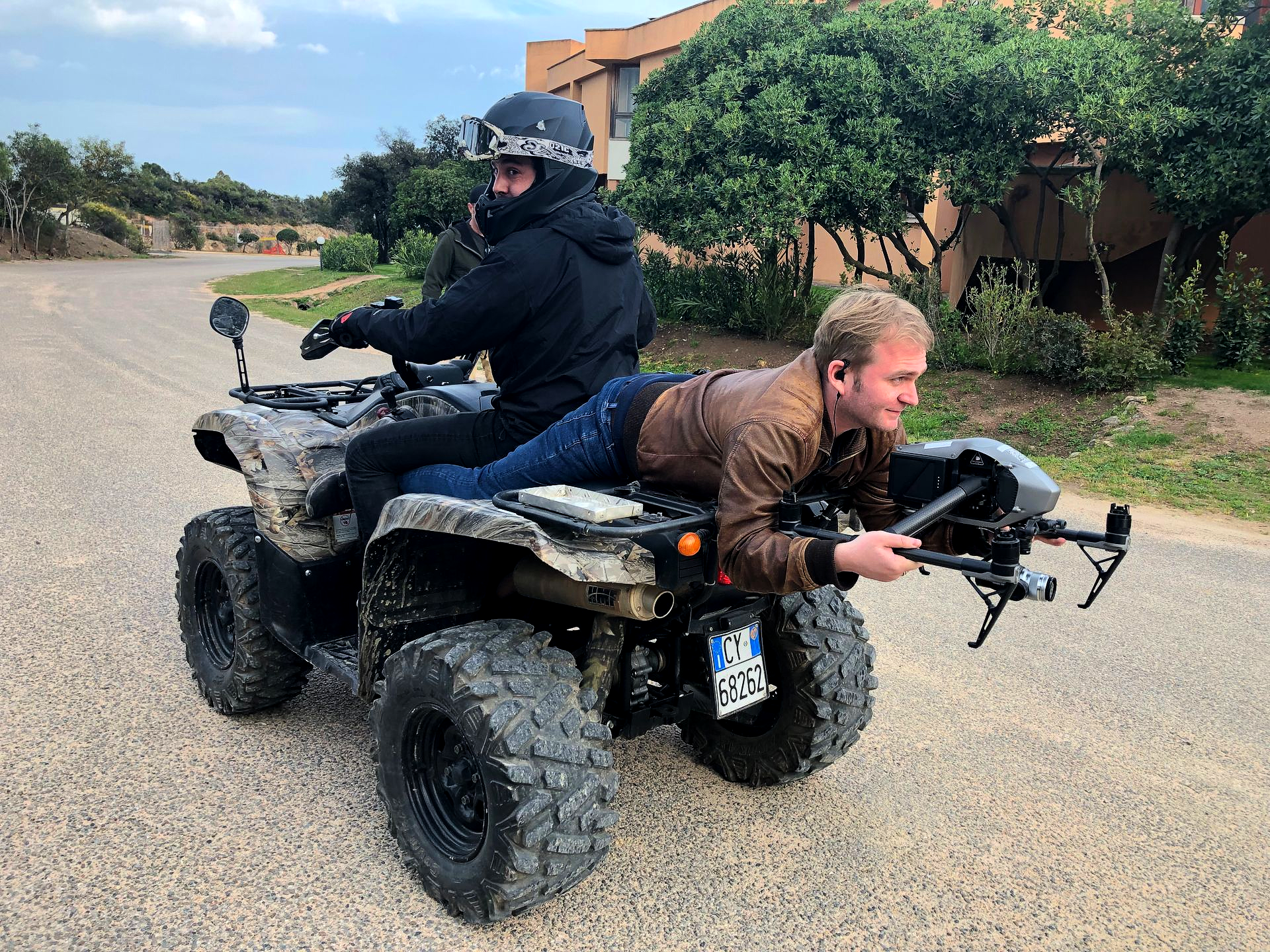 Two men on an ATV; one driving, the other aiming a device from the back. Outdoors, daytime.