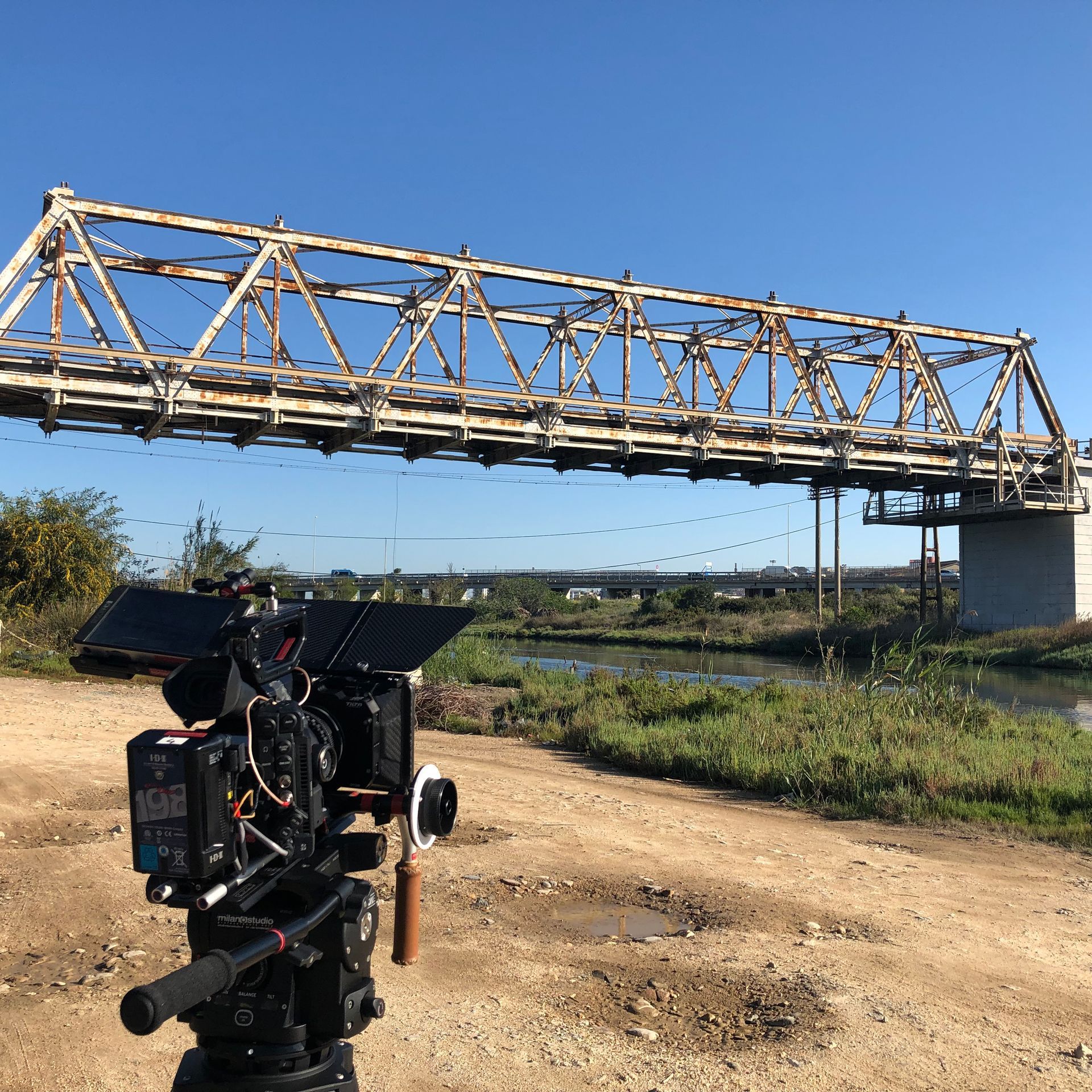 Camera on tripod, filming a weathered steel bridge over a river under a clear blue sky.