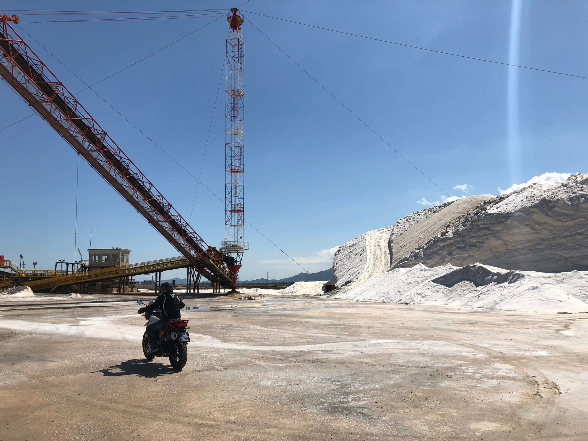 Motorcyclist rides past a large salt pile and industrial machinery under a blue sky.