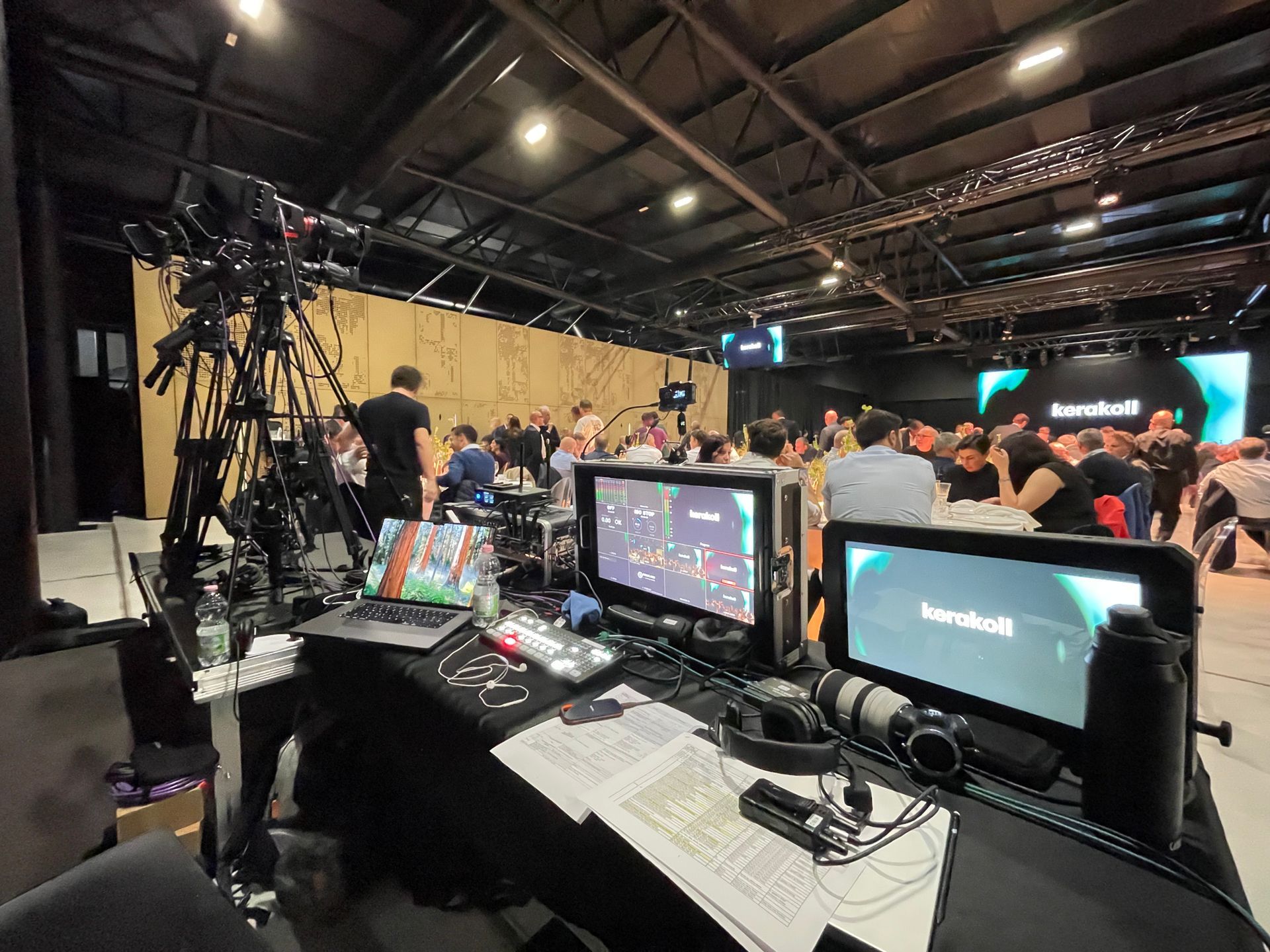 Production setup in a large hall with people. Monitors, cameras, and equipment are in the foreground.