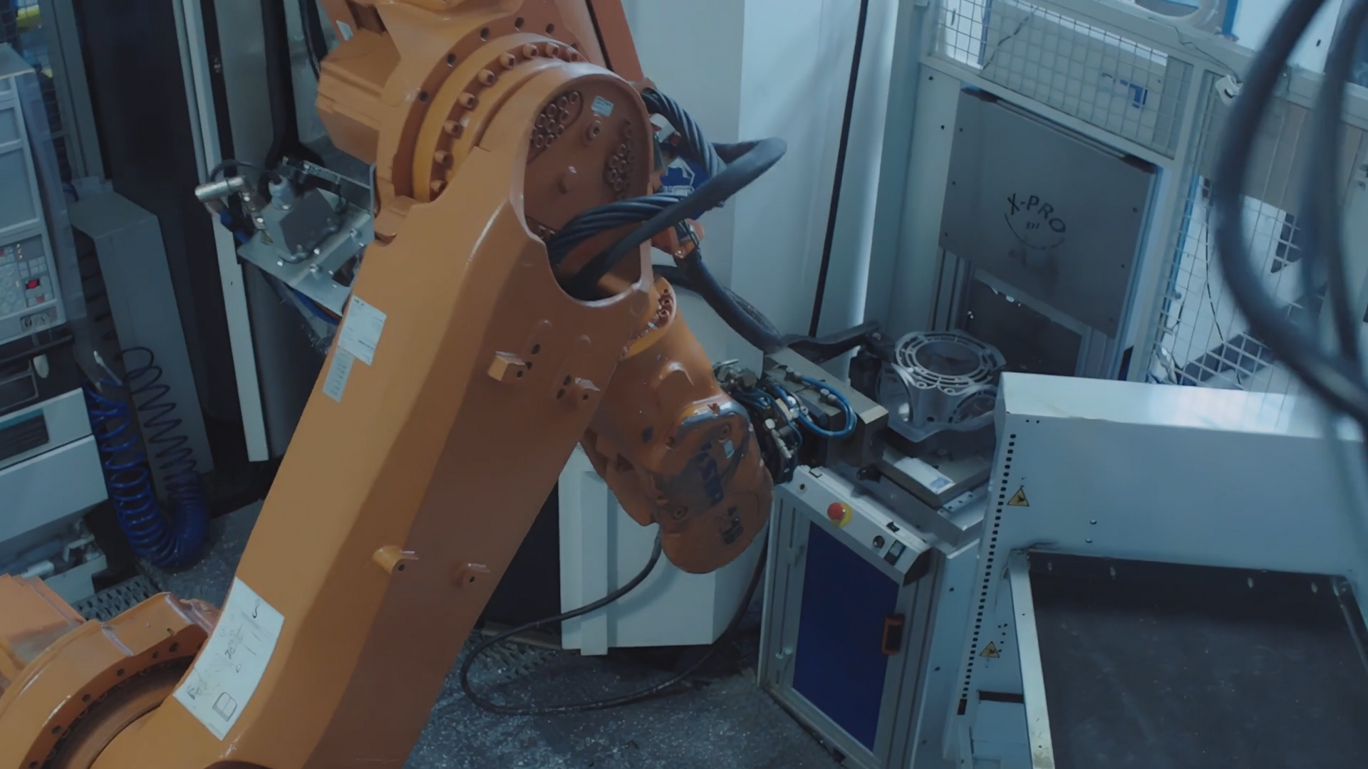 Orange robotic arm in a factory setting, manipulating an object near machinery and wires.