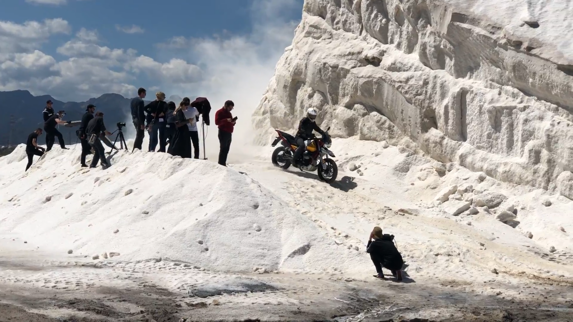 Motorcycle riding up a white, sloping hillside with several people watching on a sunny day.