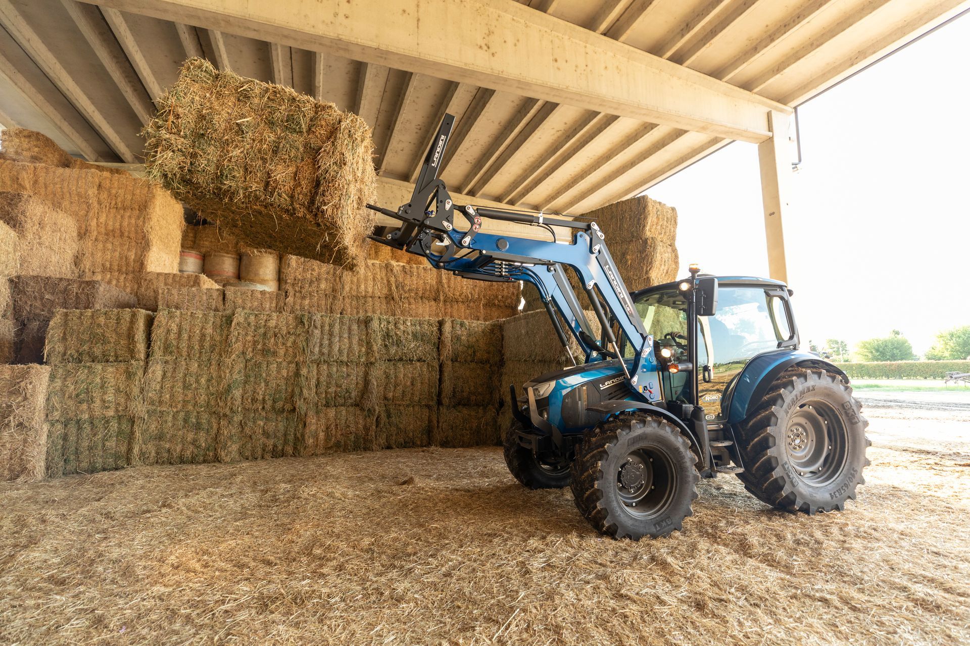 Blue tractor with hay bale on forks, stacking bales in a barn.