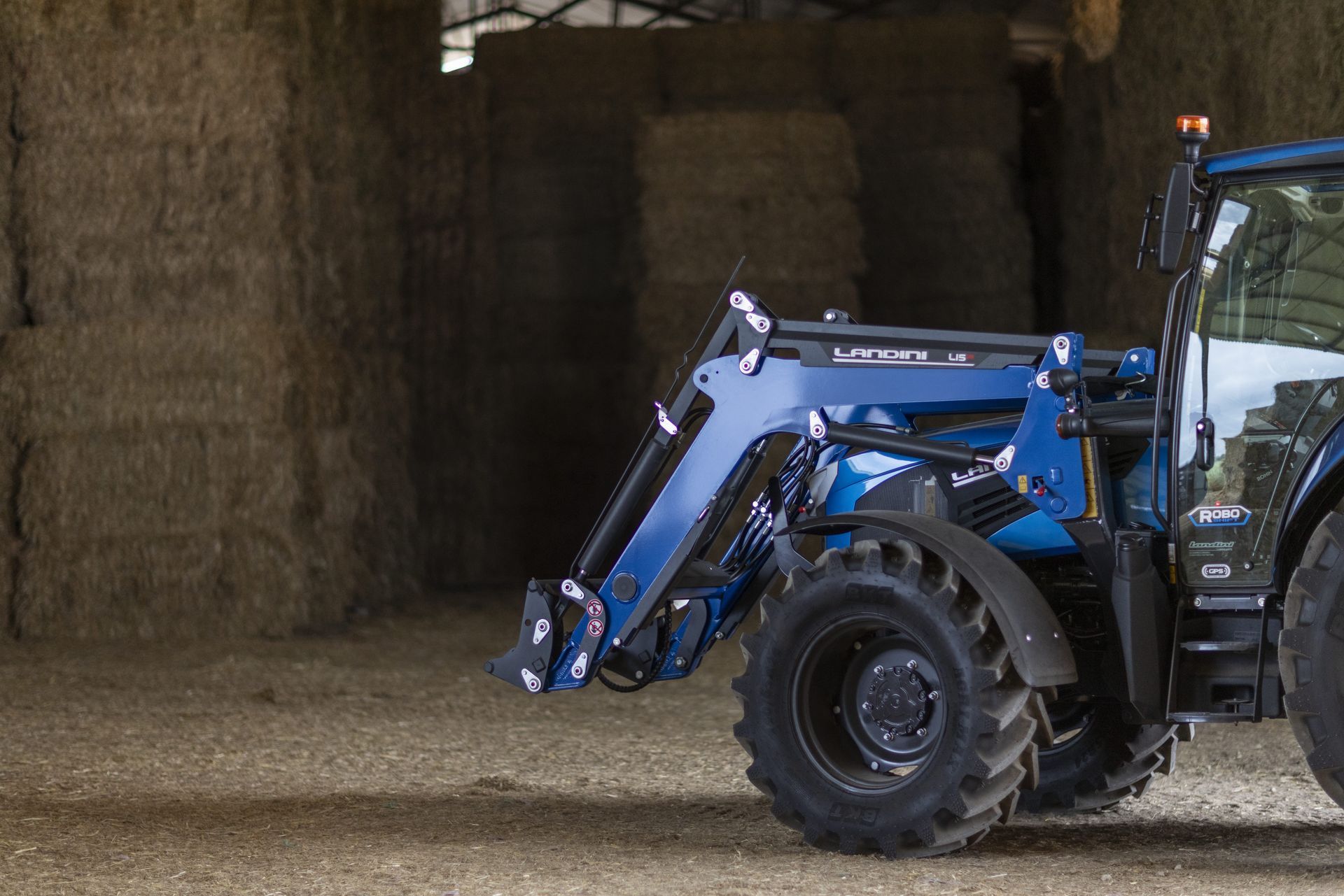 Blue tractor with front loader inside a barn with hay bales.