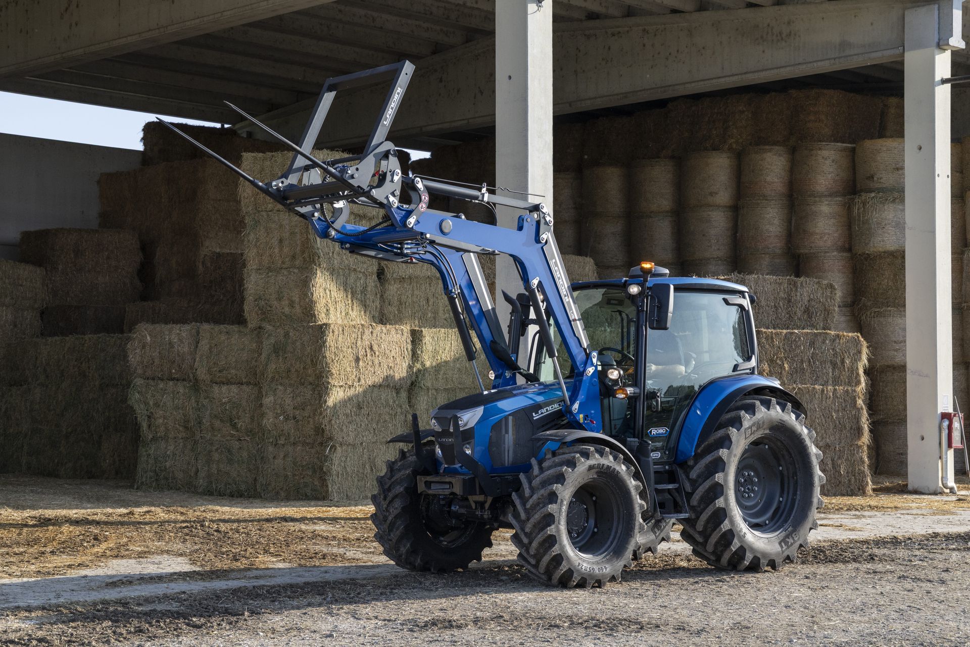 Blue tractor with hay bale loader at a farm, lifting a bale.