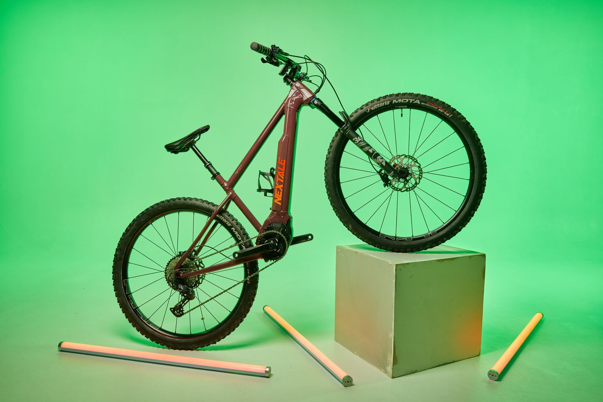 An e-bike, with one wheel on a block, set against a green backdrop. Wooden rods are in the foreground.