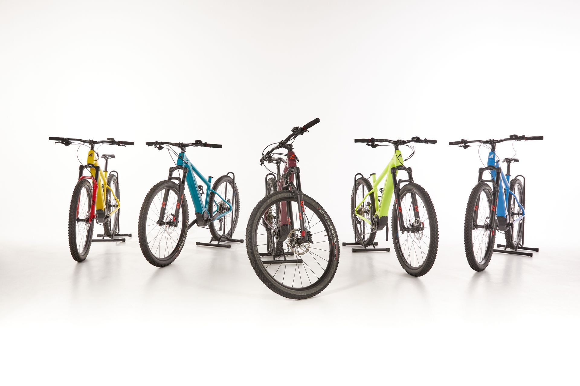 Five colorful mountain bikes on stands, arranged in a row against a white background.