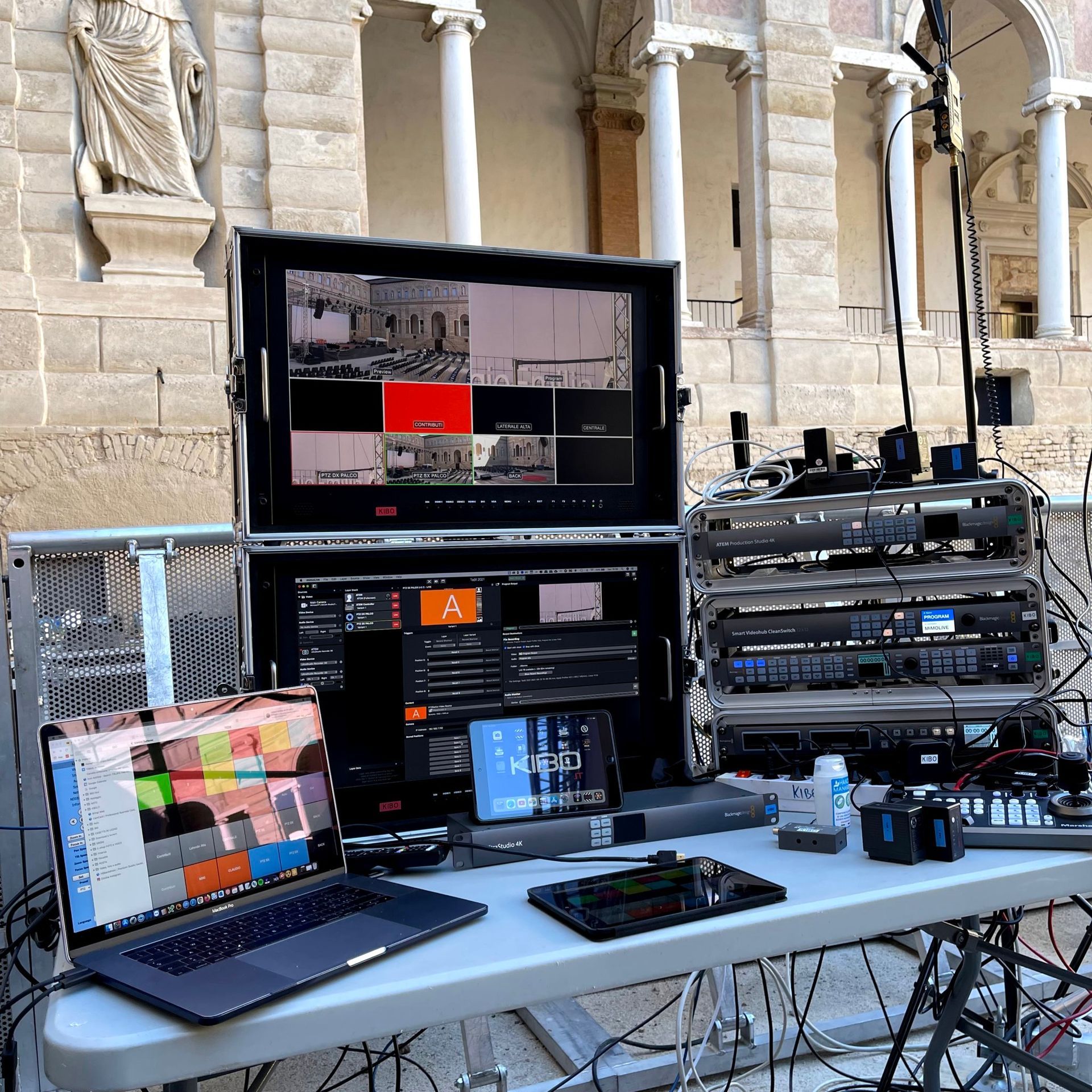 A live broadcast control station with monitors, a laptop, and audio equipment set up outdoors near a building with columns.