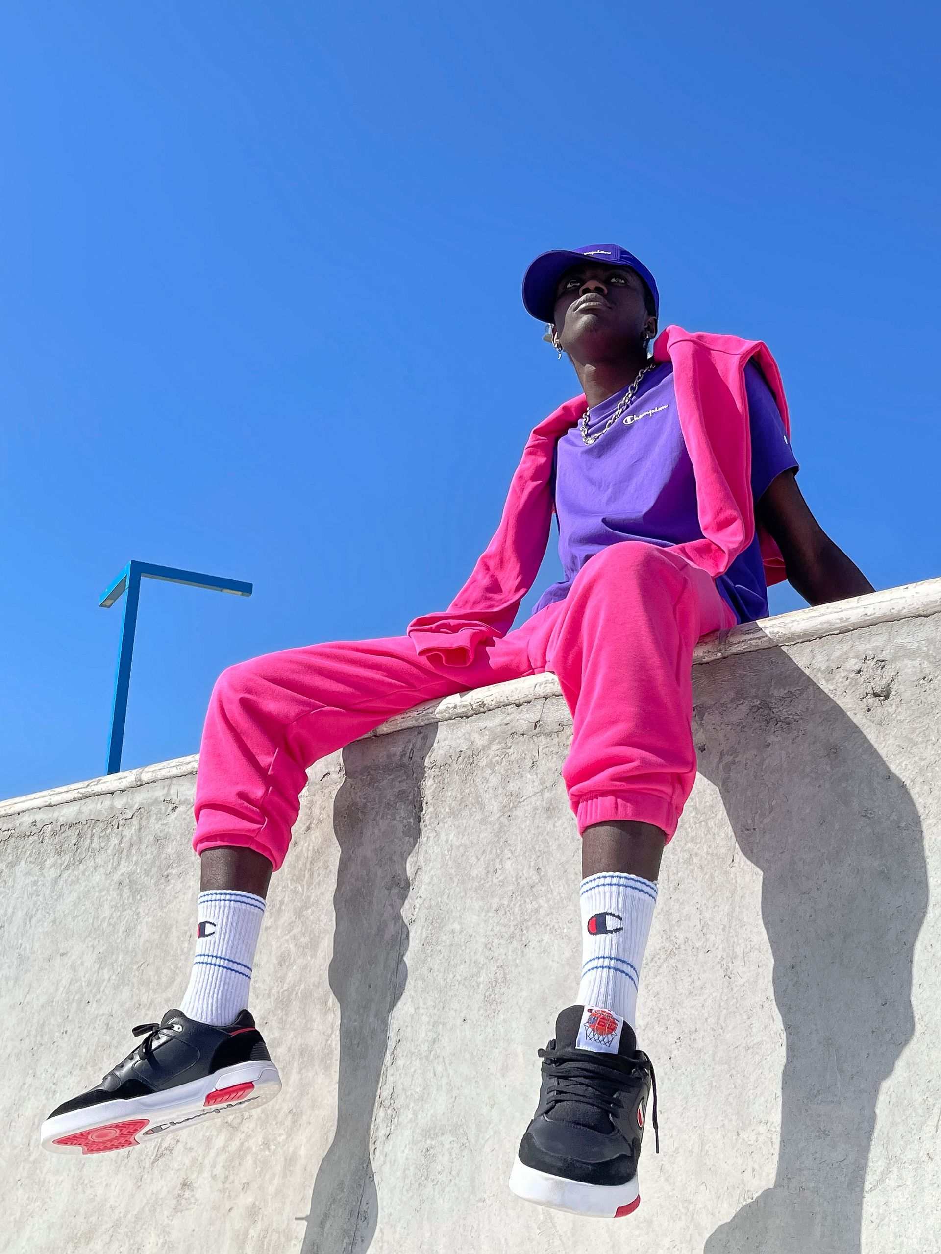 Person wearing vibrant pink and purple clothing, sitting on a wall against a blue sky.