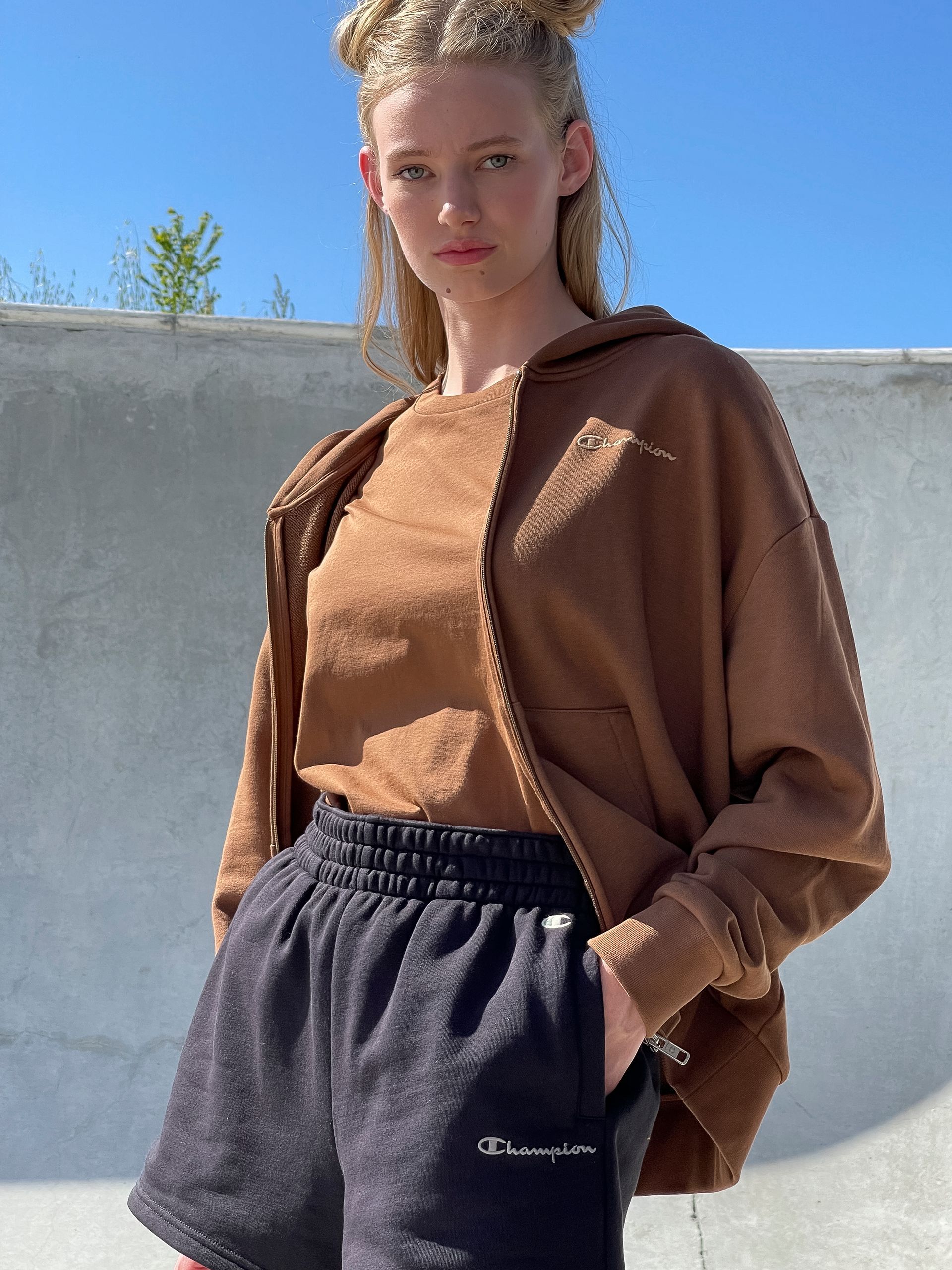 Woman in brown hoodie, tee, and black shorts stands with hands in pockets. Outdoors, against a concrete wall.