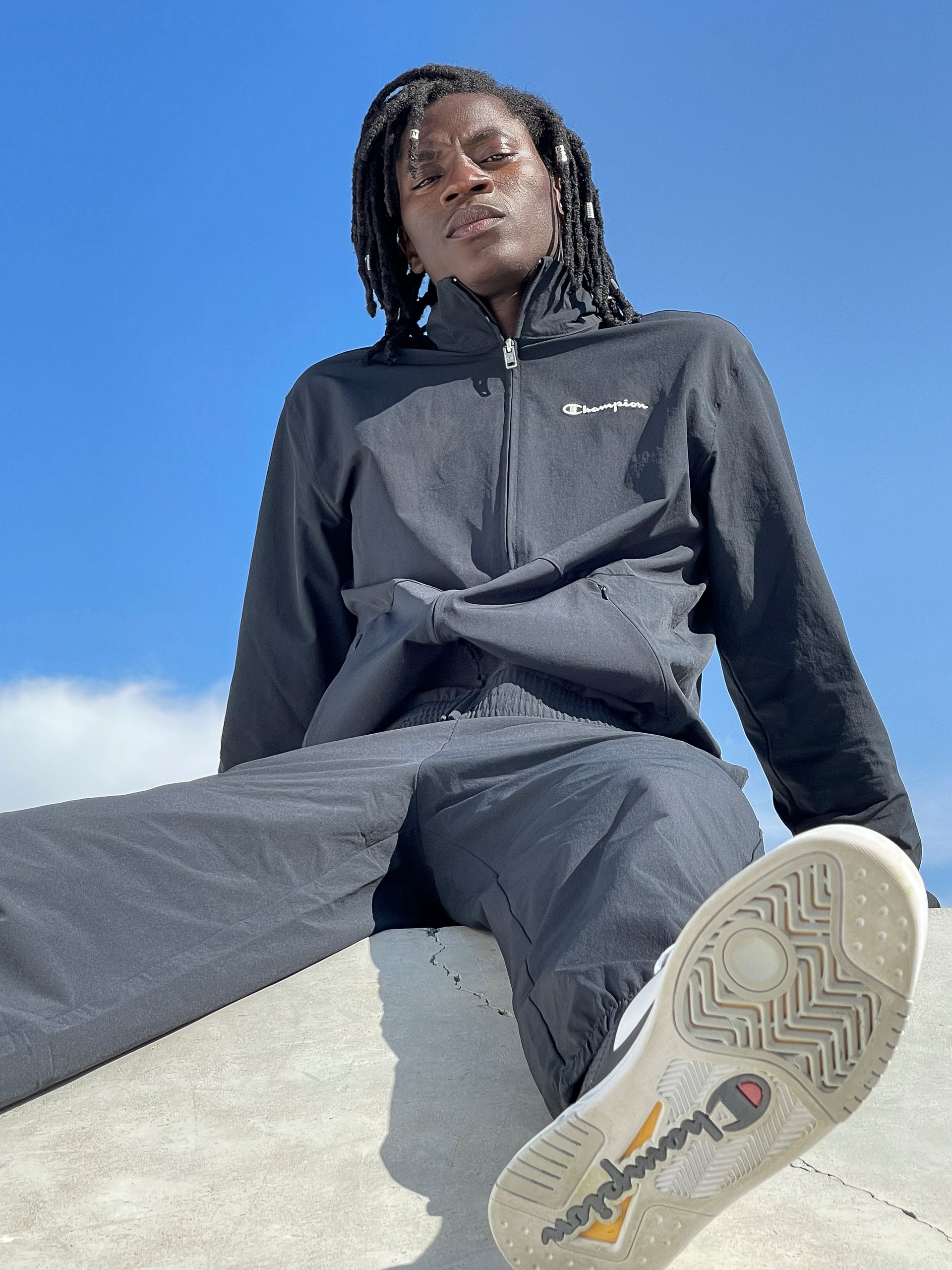 Person in a black Champion tracksuit, sitting, against a blue sky, leg extended, shoe sole visible.