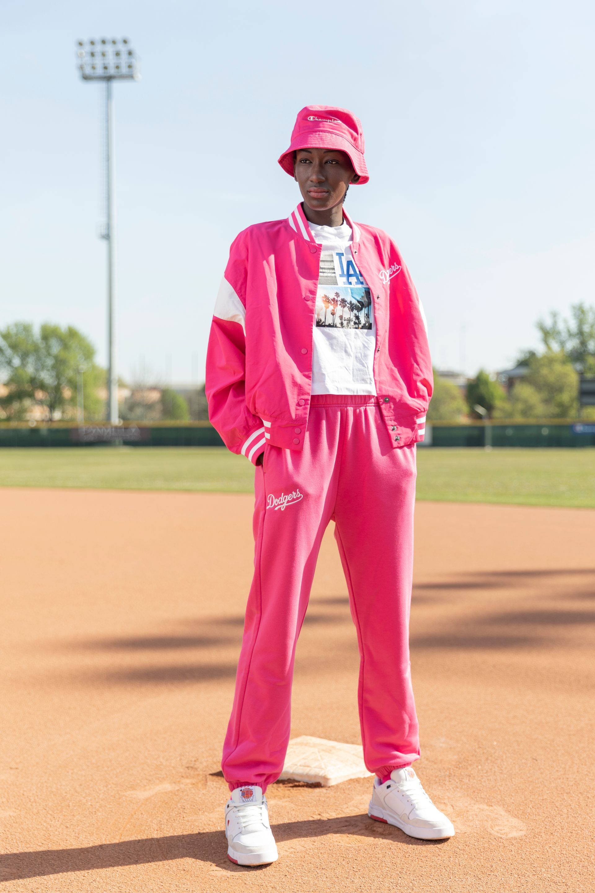 Person in pink tracksuit and bucket hat stands on a baseball field.