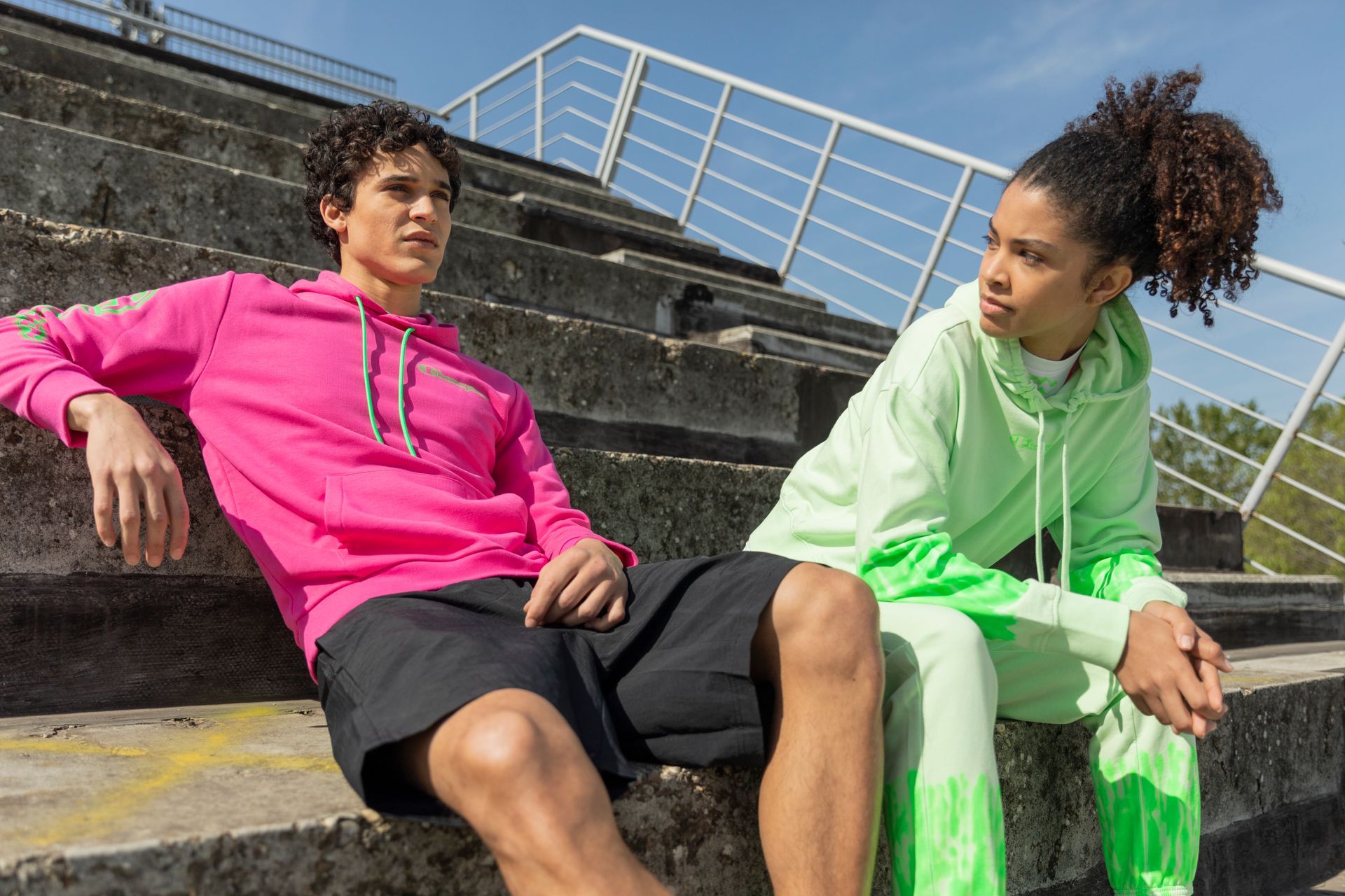 Two people in bright hoodies sit on stadium steps, looking thoughtful.