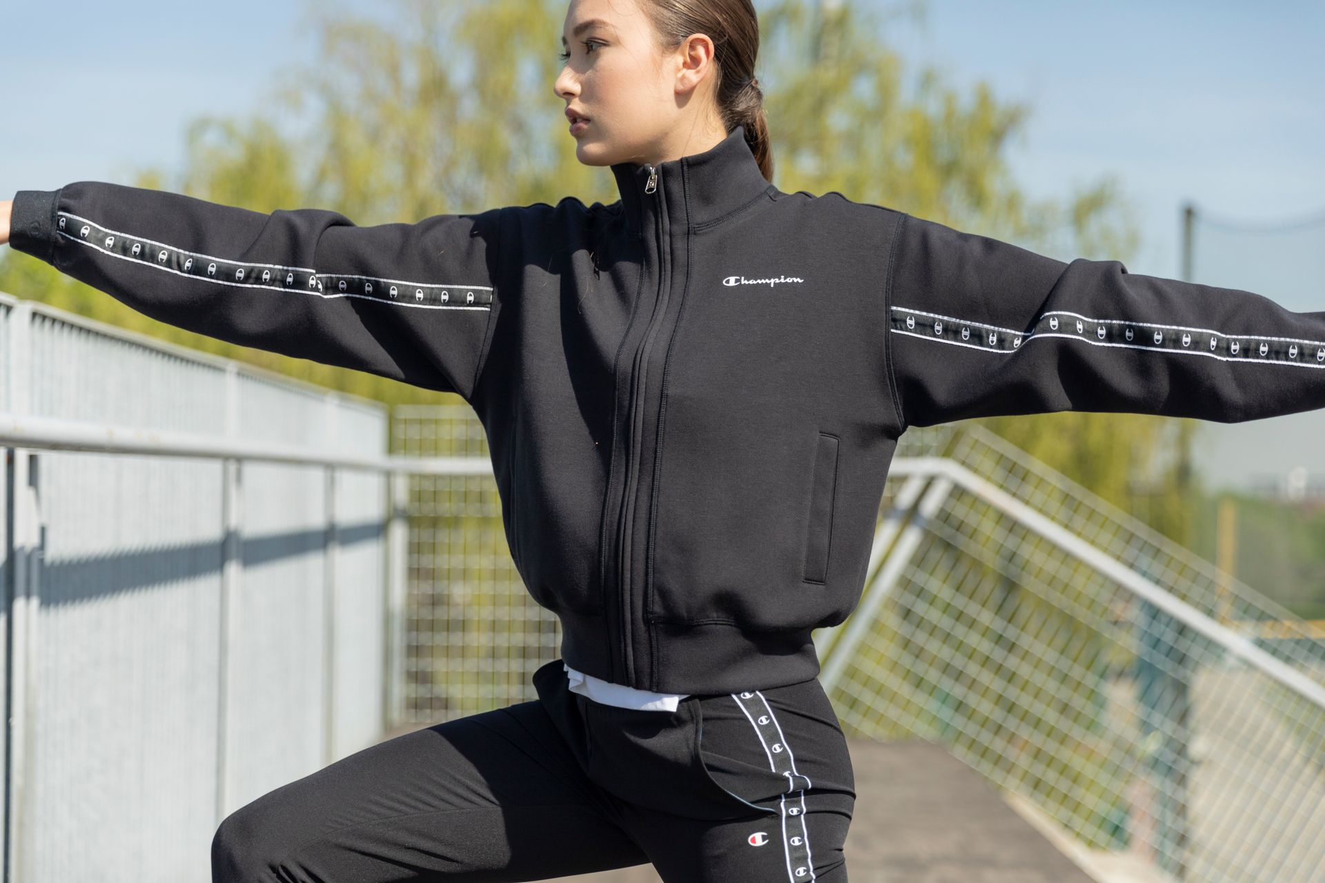 Woman in black Champion tracksuit, arms outstretched, near bleachers outdoors.