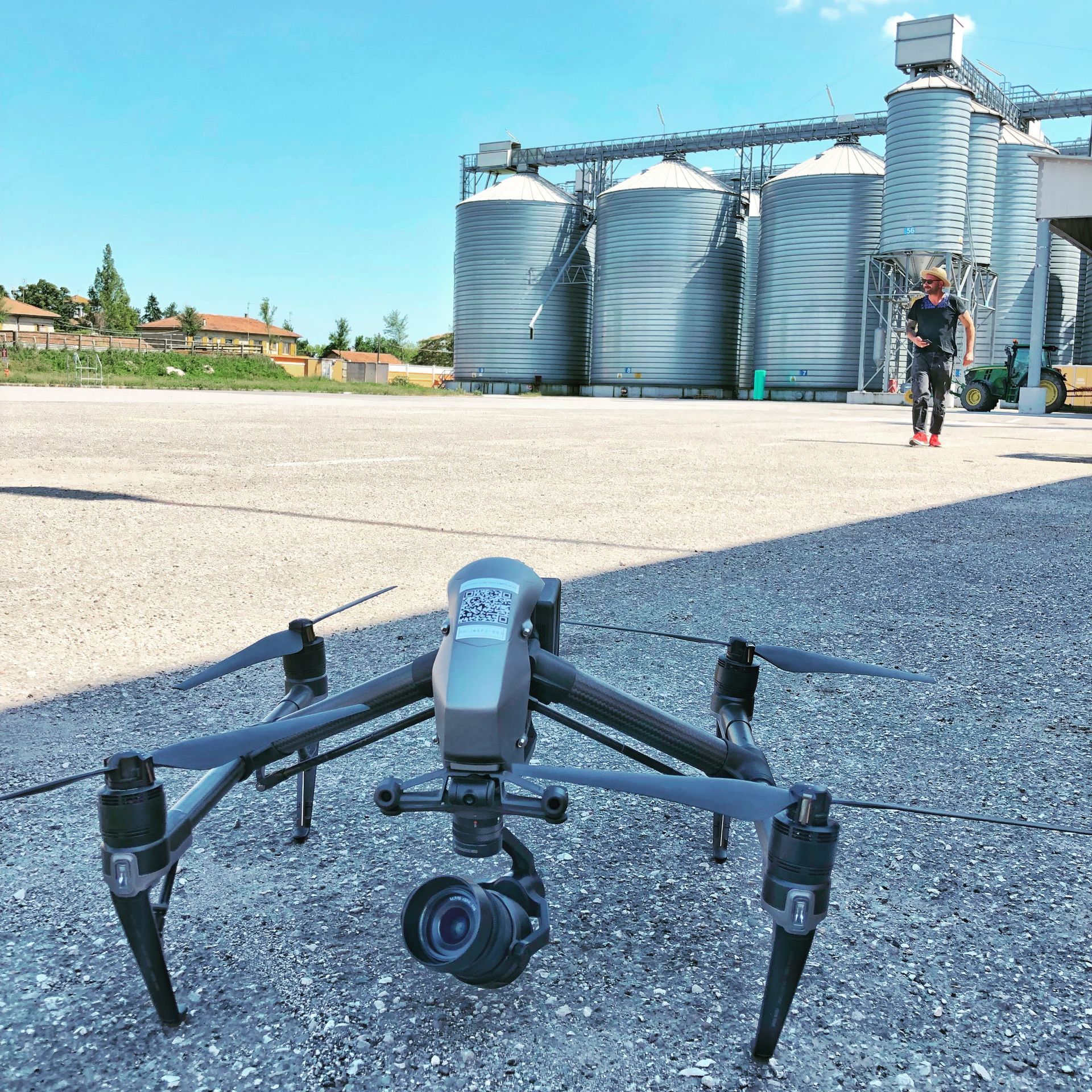 Drone with camera on the ground, man in distance by grain silos on a sunny day.