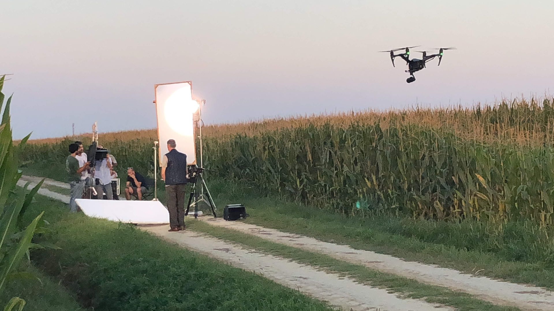 A drone flies over a film set in a cornfield. A person stands near a large light reflector, with crew members.