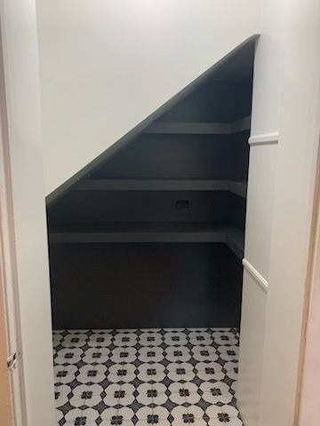 Storage closet under stairs with black shelves, gray walls, and patterned floor.