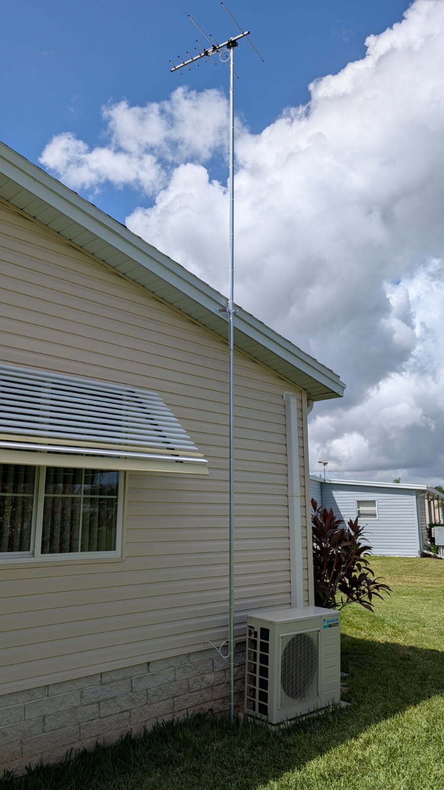 Television antenna mounted on a tall pole beside a beige house. Blue sky and clouds in the background.