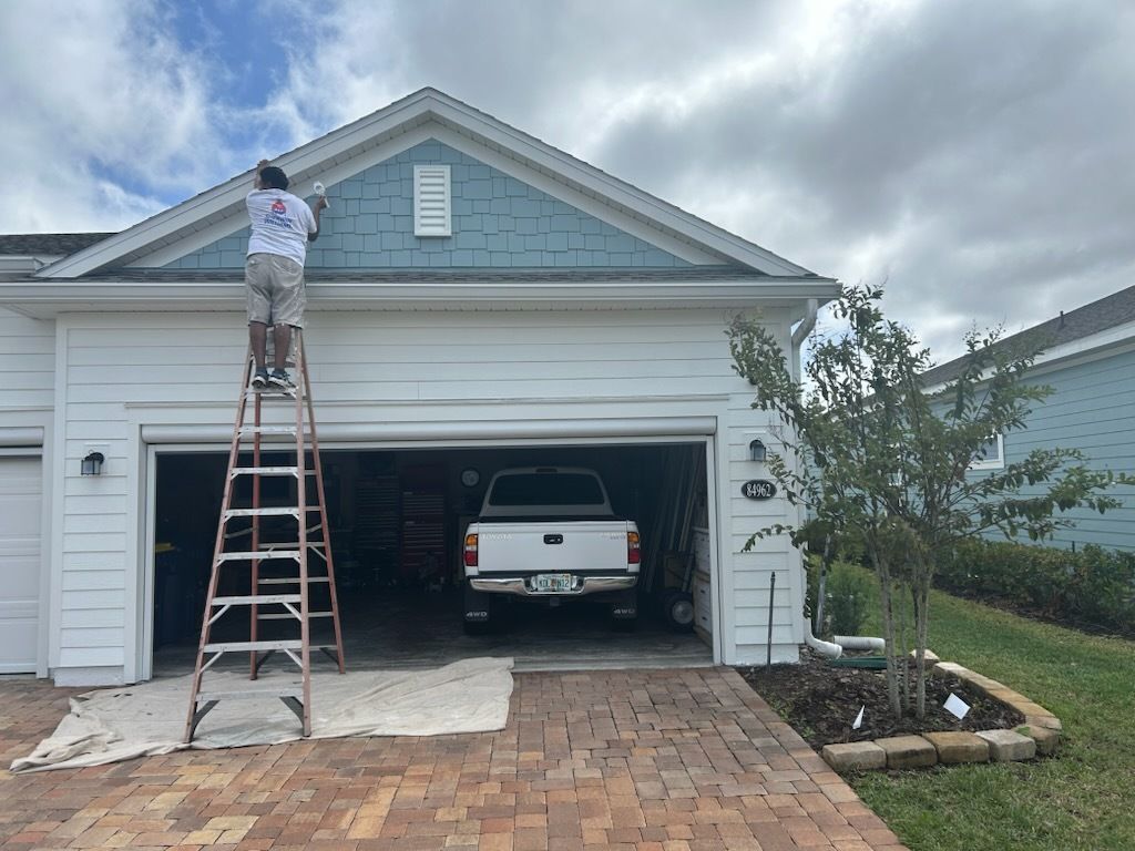 Man on ladder paints a house. The garage door is open, revealing a truck inside. Cloudy sky.