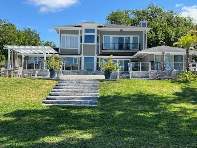 Large waterfront house with steps leading to the entrance. Green grass, blue sky, and a pergola are present.