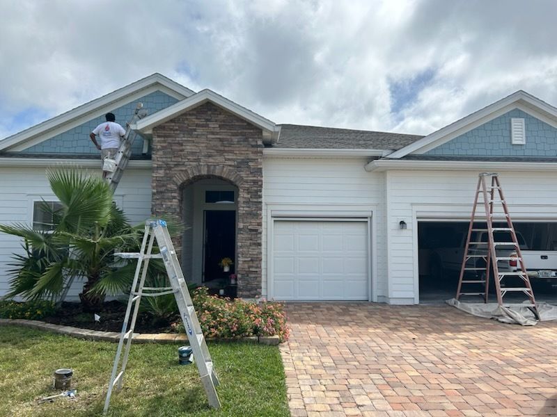 Exterior house painting: A worker on a ladder paints white siding with blue accents; brick driveway.