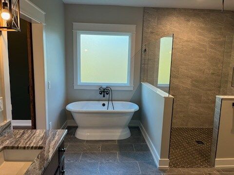 Freestanding tub under a window in a modern bathroom with stone tile, adjacent shower, and granite countertop.