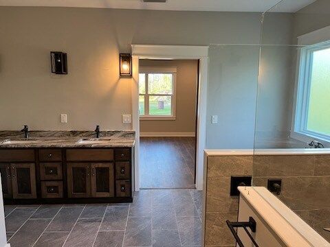 Bathroom with a double vanity, gray tile floor, and a doorway to another room with a window.