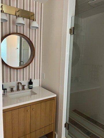 Bathroom with a wooden vanity, round mirror, striped wallpaper, and a glass shower door.