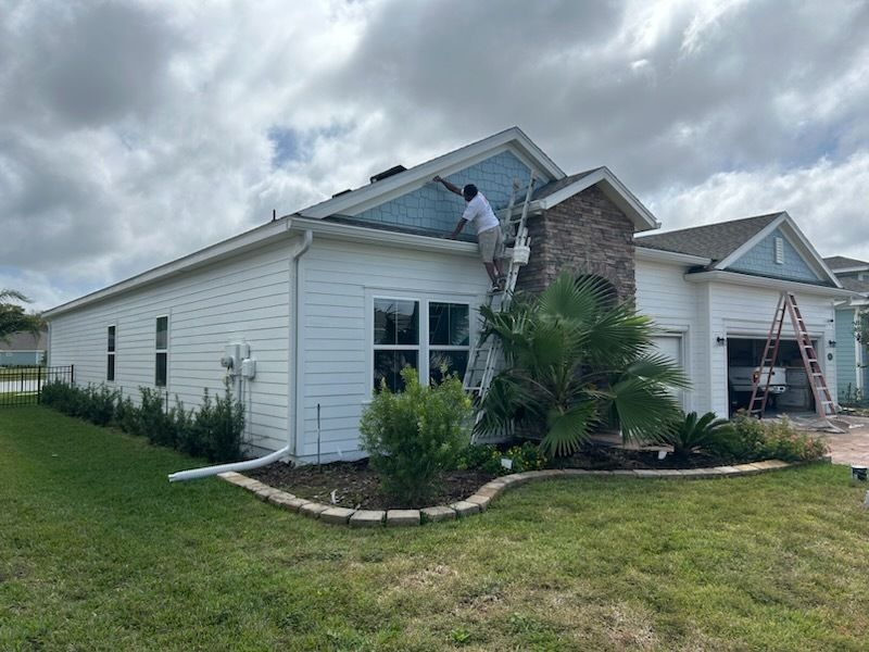 Person painting a white house with blue trim; ladder, cloudy sky, green grass.
