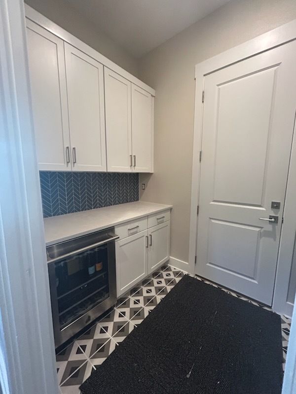 Pantry with white cabinets, blue backsplash, wine fridge, and patterned floor. Door on the right.