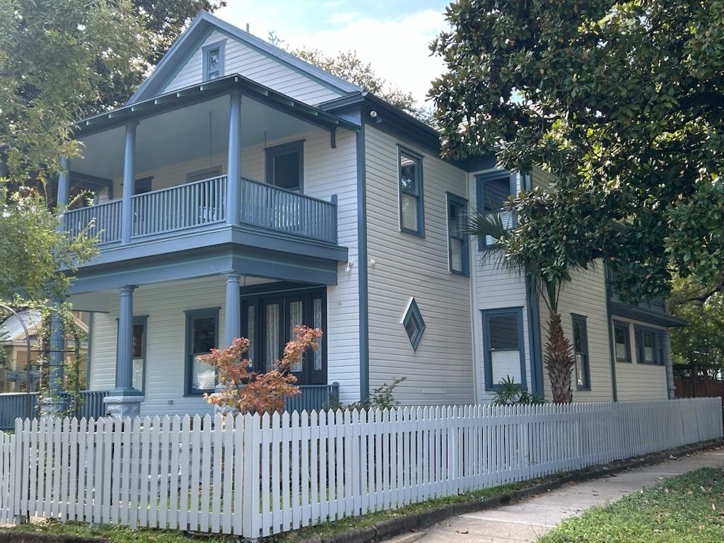 Two-story white house with blue trim, balcony, picket fence, trees.