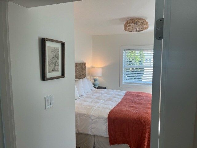 Bedroom interior: bed with white linens, orange throw, art on the wall, and a window with blinds.