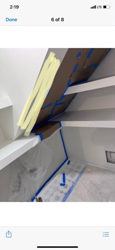 Interior view of a built-in shelf unit being painted. Blue tape, plastic sheeting, and yellow masking tape are visible.