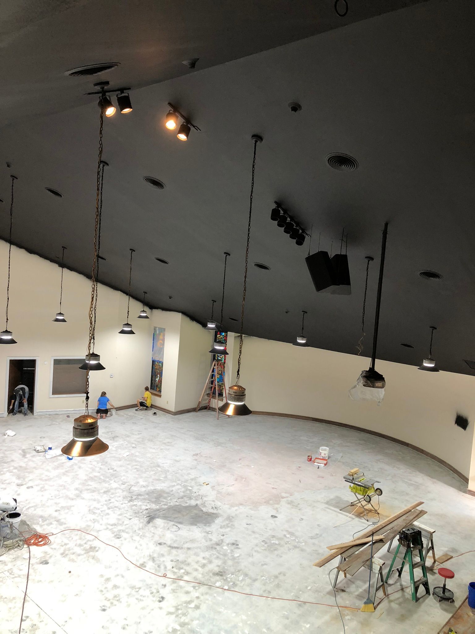 Construction in a large, curved room with a black ceiling, spotlights, and hanging lights; workers on the floor.