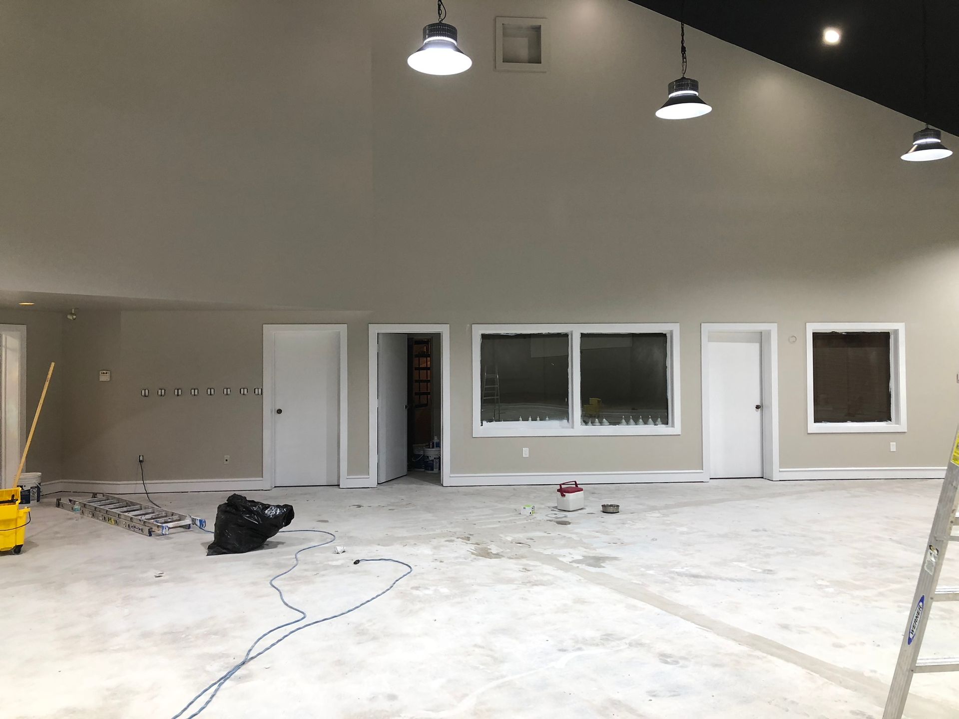 Interior construction site with neutral walls, concrete floor, windows, doors, and hanging lights.