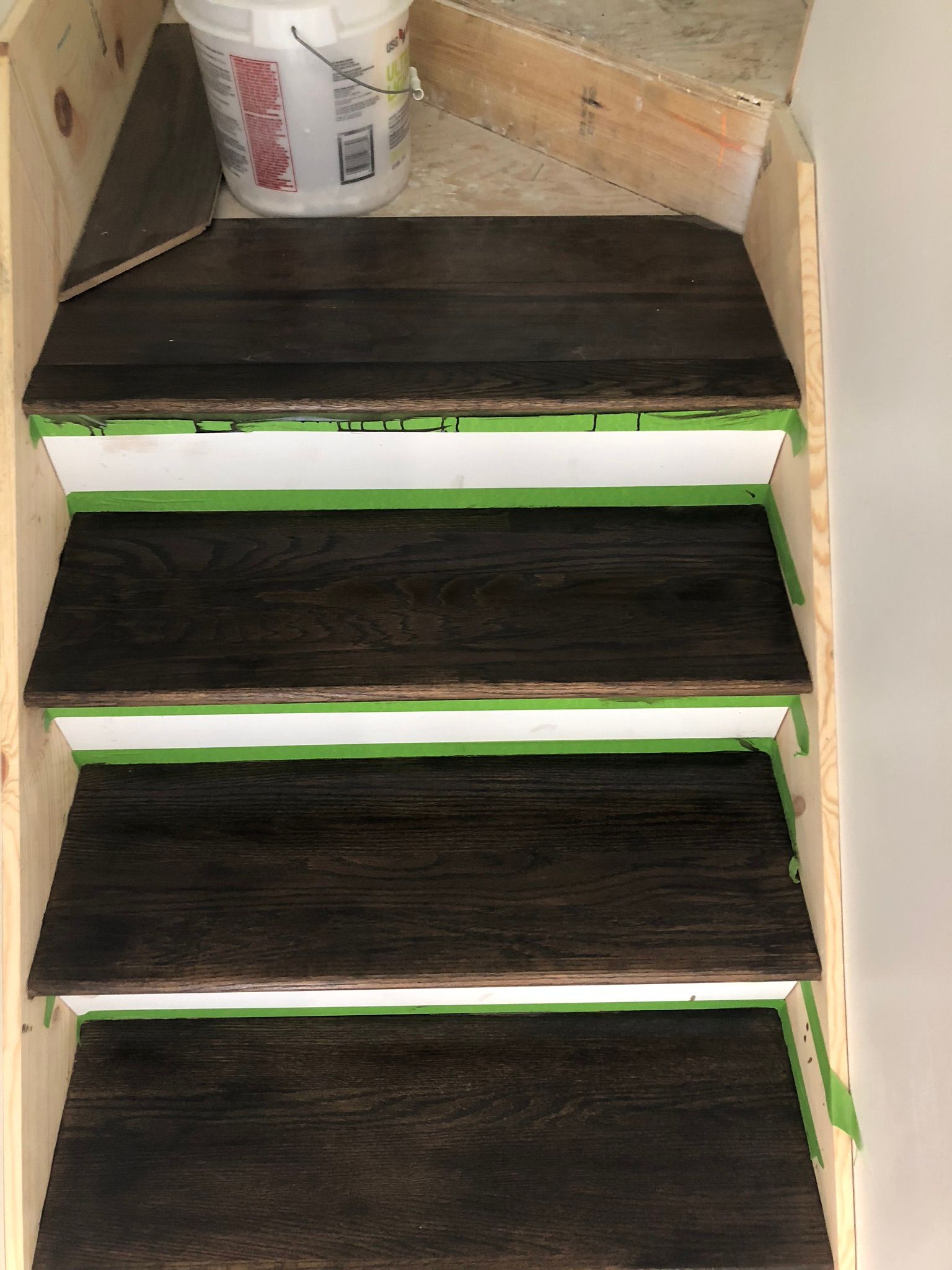 Staircase with dark wood treads, white risers, and green tape; a bucket sits on the top step.
