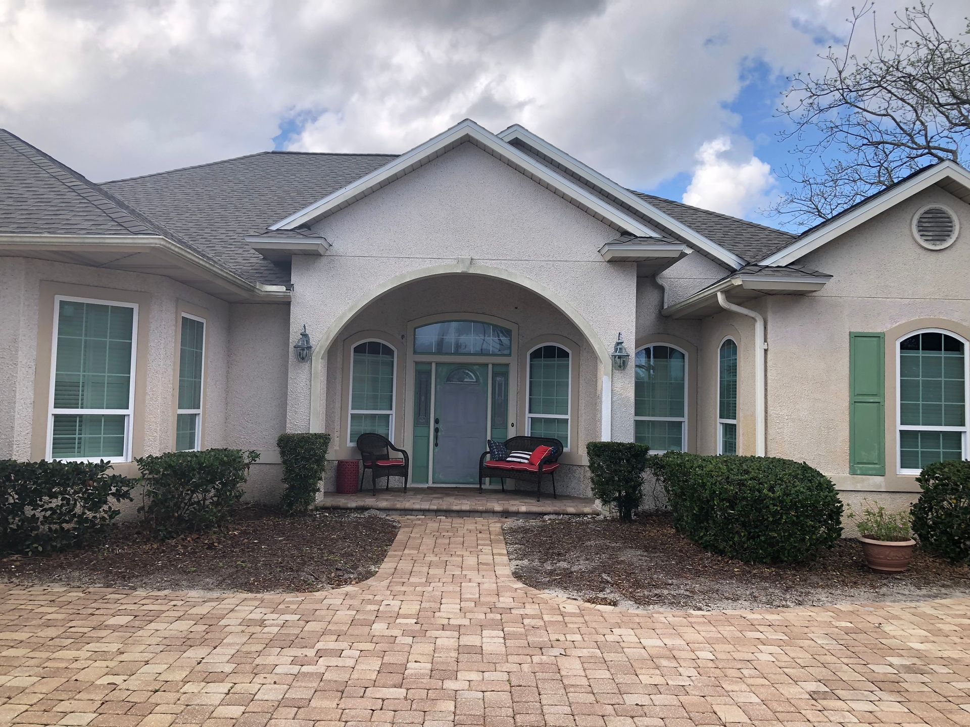 Beige stucco house with brick walkway, arched doorway, and green shutters.