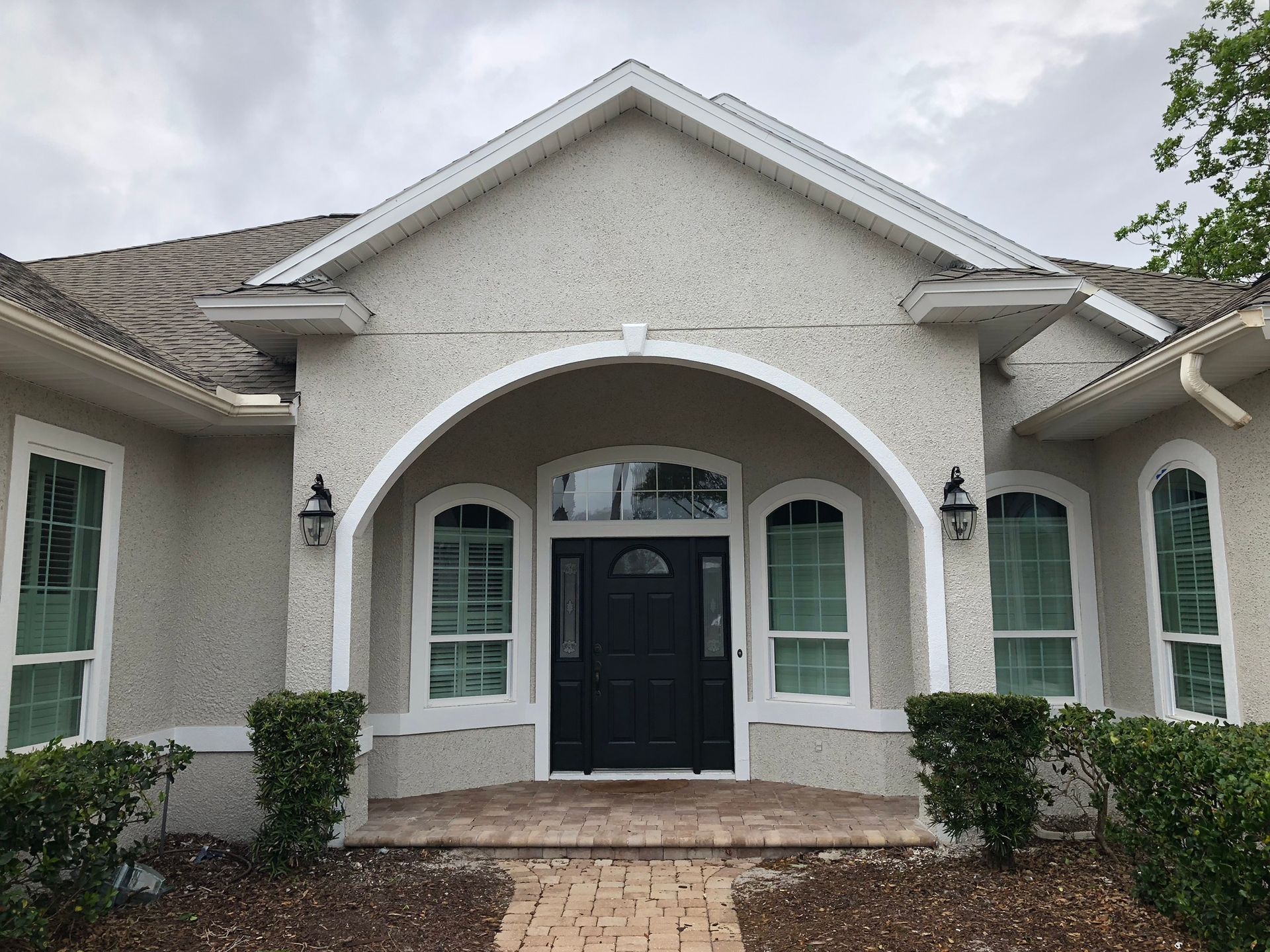 Front view of a light-colored stucco house with an arched entry, black front door, and green bushes.