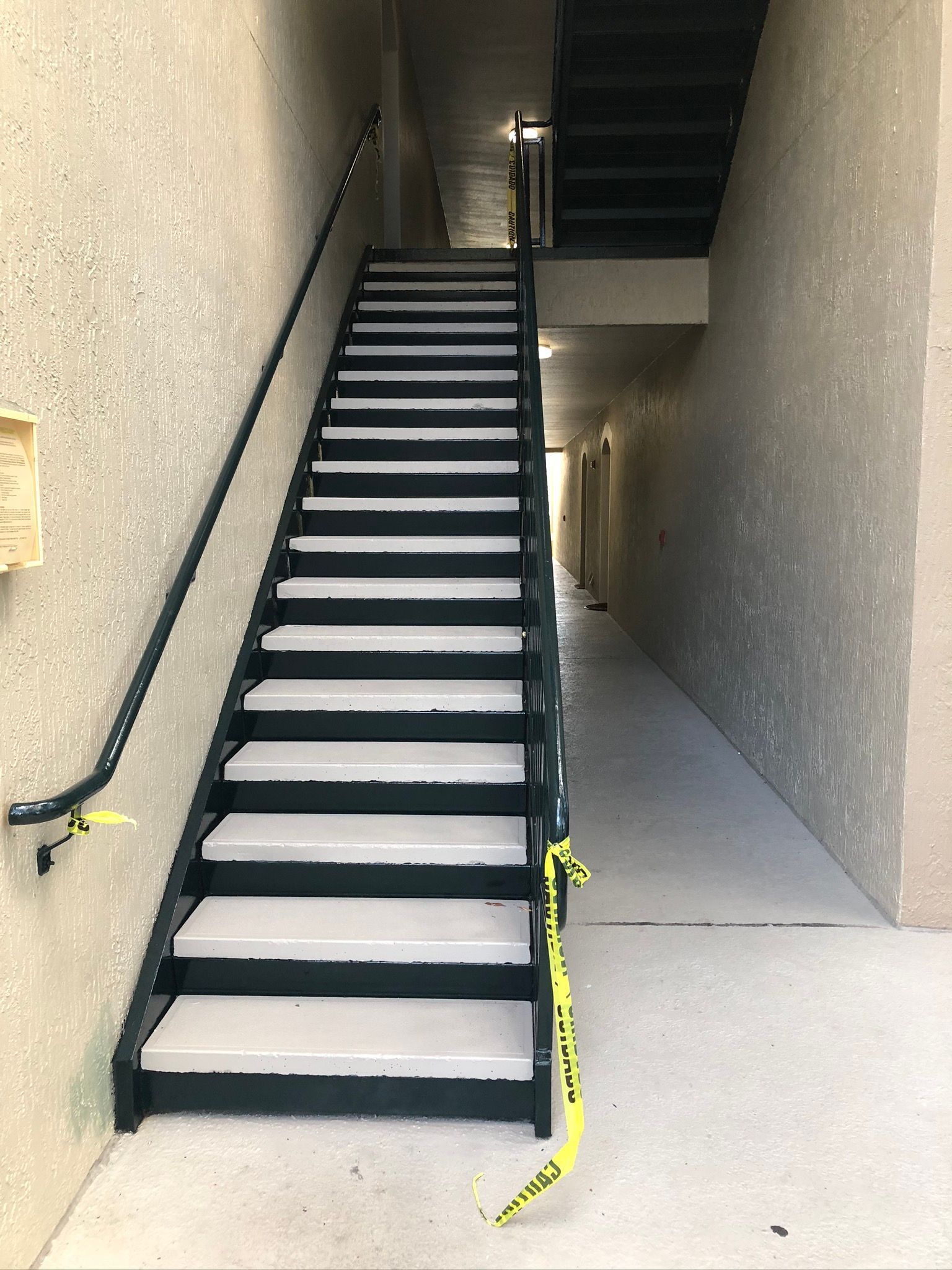 Staircase with black handrail and white steps, yellow caution tape, in a beige building hallway.