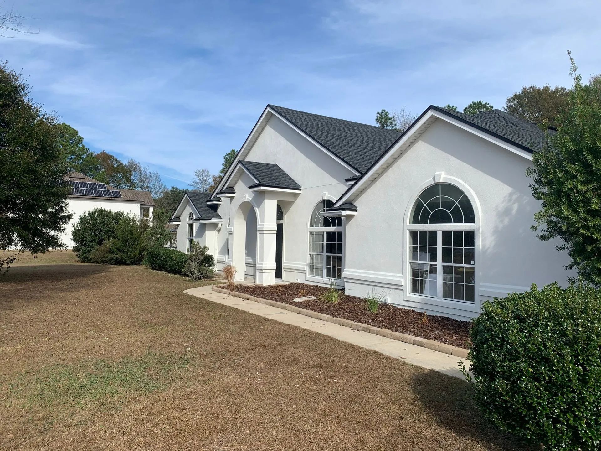 White suburban house with arched windows, front walkway, and a large brown lawn under a blue sky