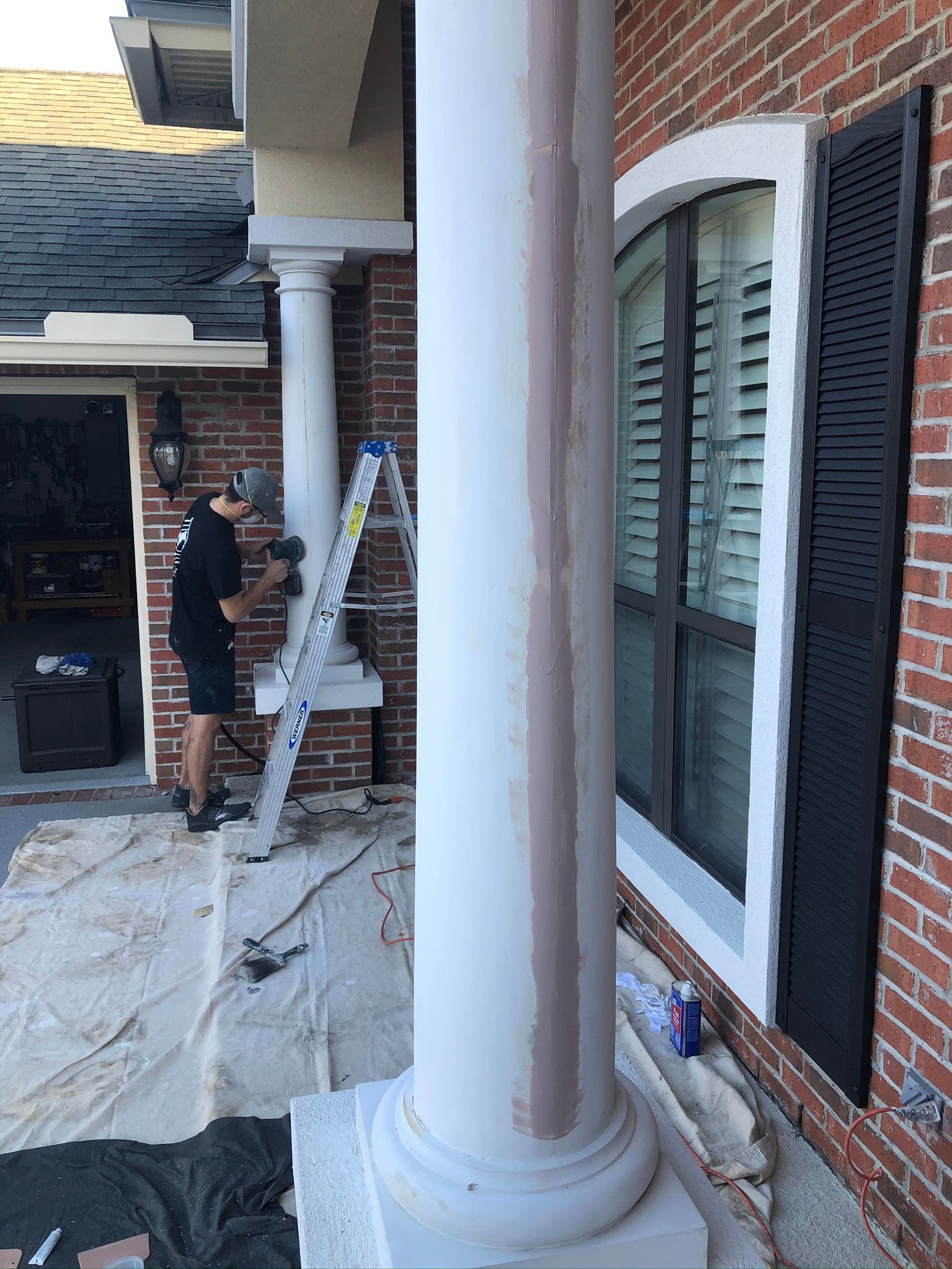 Man sanding a white pillar next to a brick building with a window.