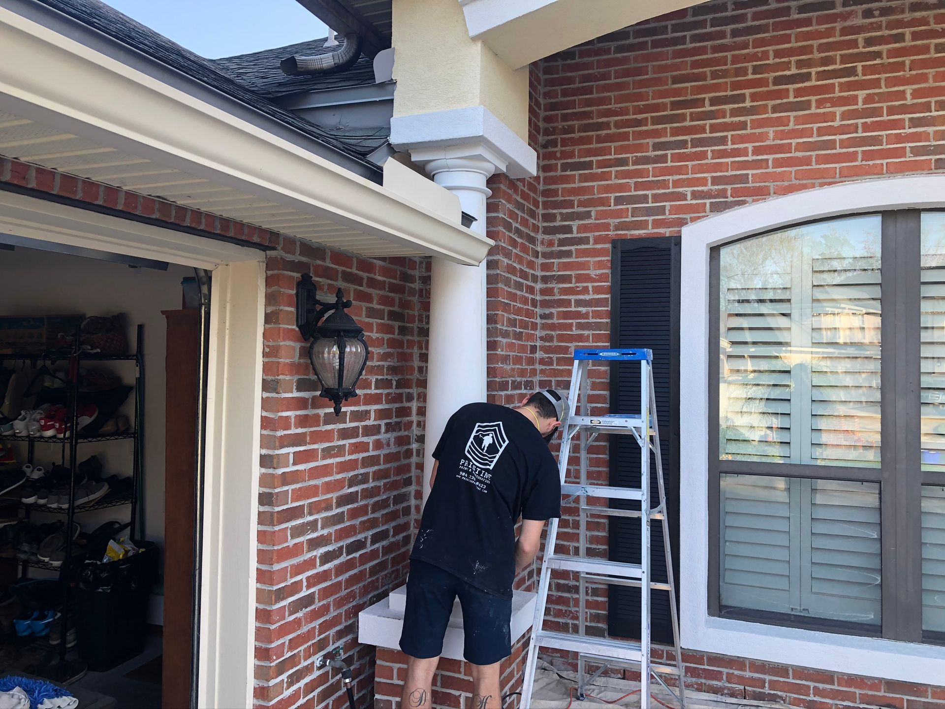 Man on a ladder painting the exterior of a brick house, near a window and garage.