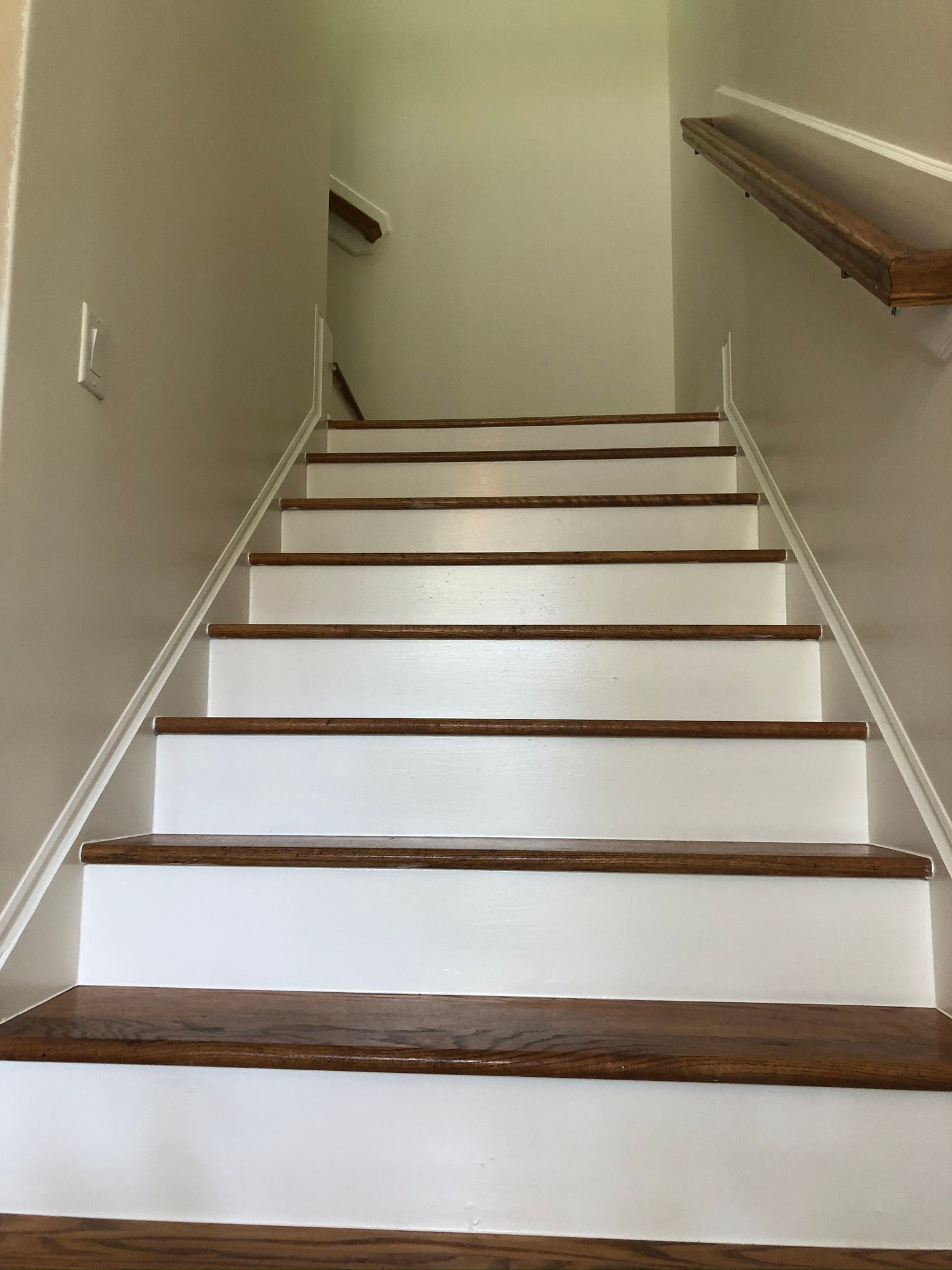 Staircase with brown wooden steps, white risers, and a wooden handrail against a cream-colored wall.