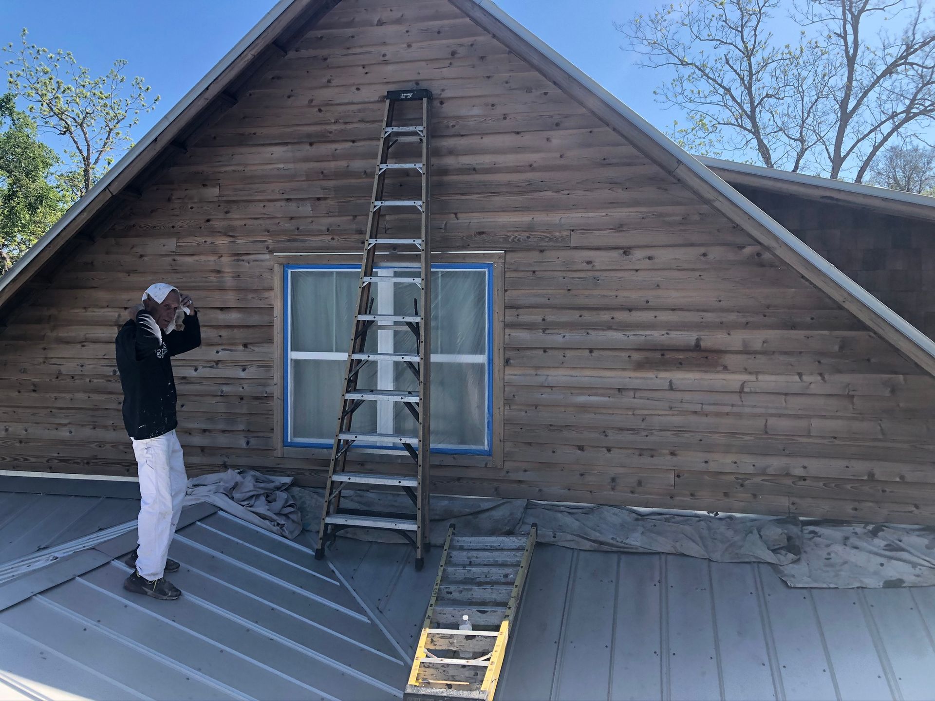 Man on roof with ladder leaning against wood-sided house, prepping for painting. Blue tape on window.