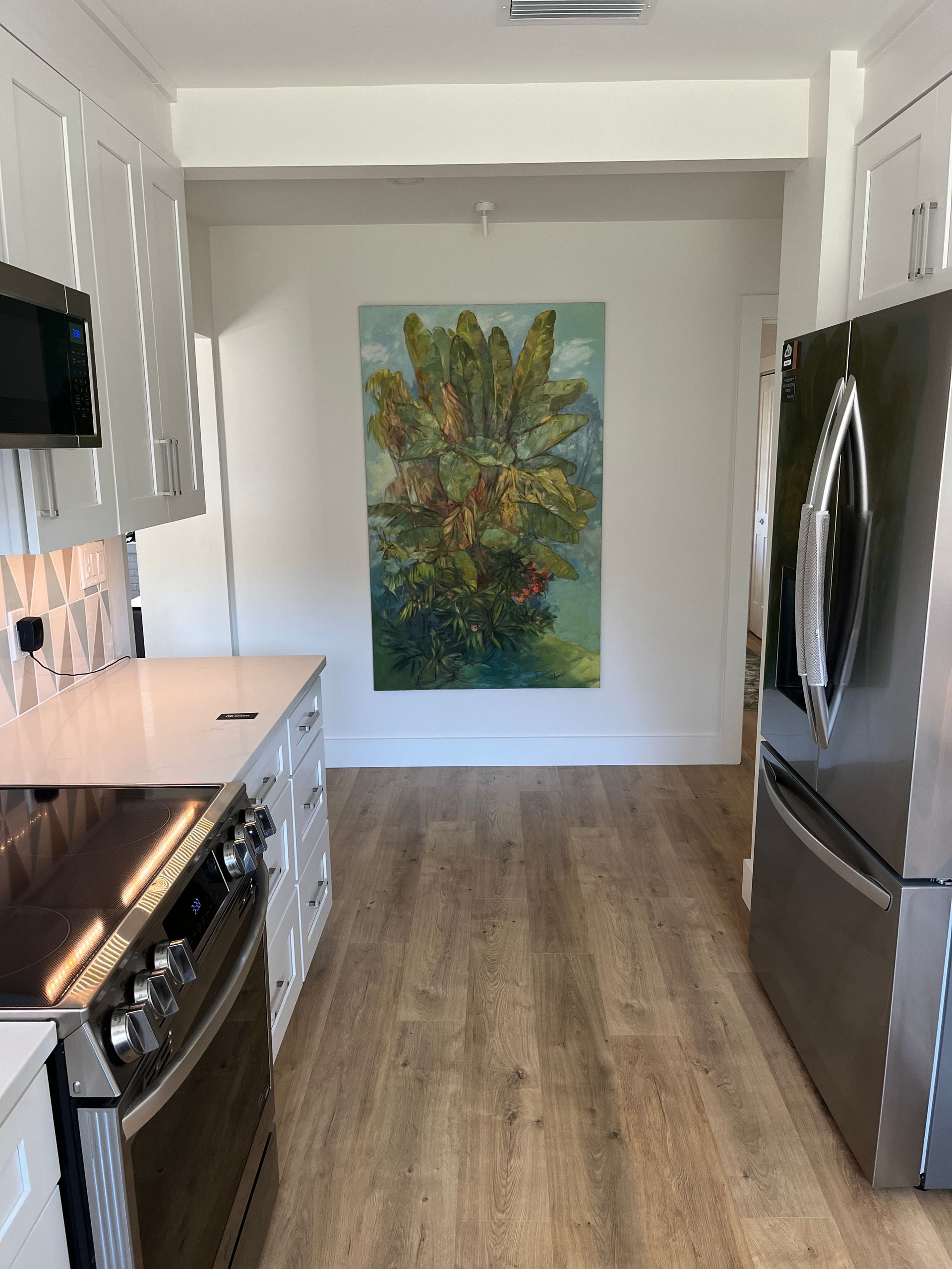 Kitchen with white cabinets, appliances, light wood floor, and a painting of a tree.
