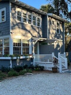 Two-story blue house with white trim and porch, nestled amongst greenery. Gravel driveway in front.