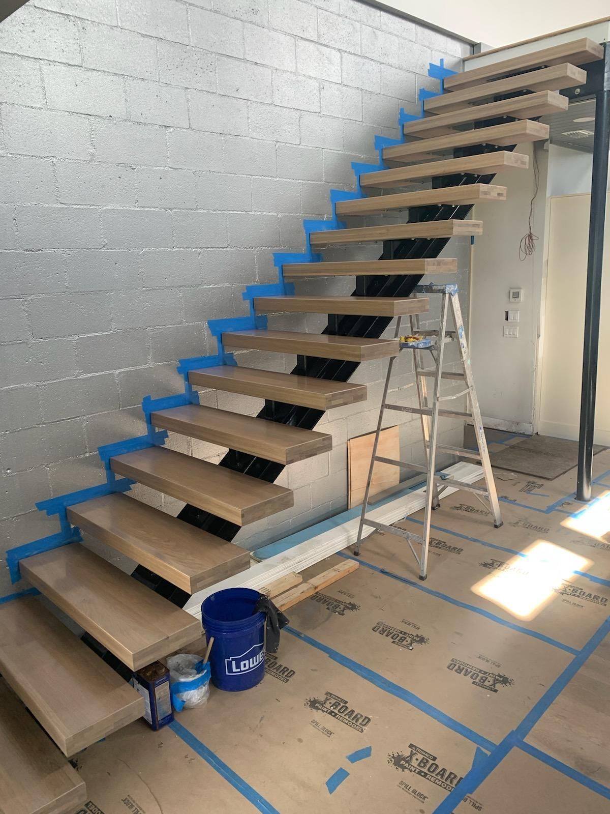 A wooden staircase is being painted blue in a room with a ladder.