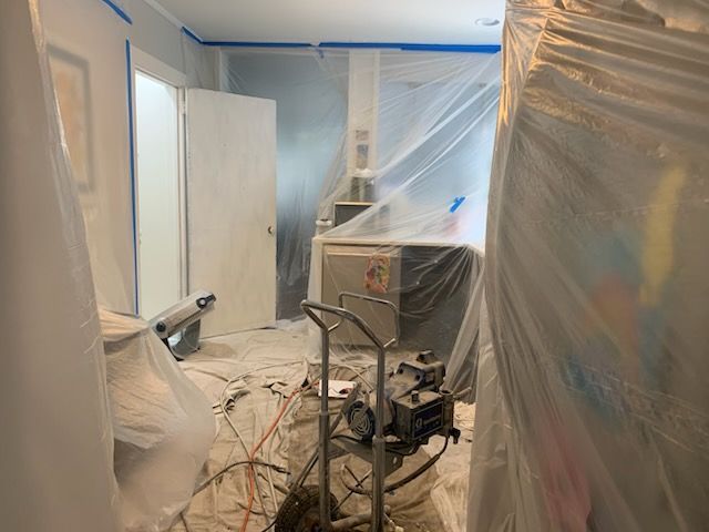 Room being painted, covered in plastic sheeting. Paint sprayer and equipment are visible.