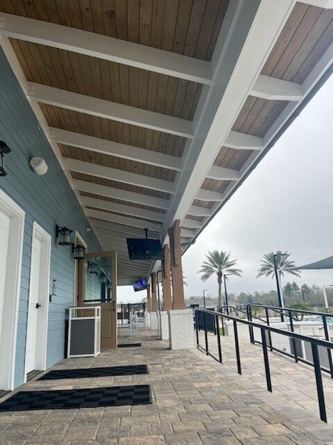 Outdoor covered walkway with blue wall, wood ceiling, and brick pavers. Palm trees visible in the distance.