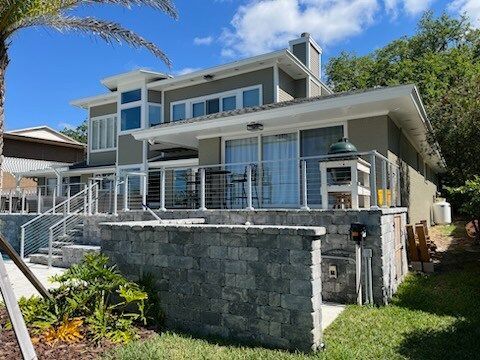 Two-story house with a deck, light gray exterior, and a stone retaining wall. Blue sky.